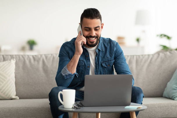 Werk vanuit huis. Positieve jonge Arabische man praten op mobiele telefoon tijdens het gebruik van laptop computer binnen - Foto, afbeelding