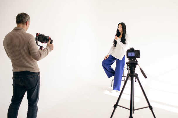 Uma jovem blogueira filma um vídeo blog no estúdio com a ajuda de um videógrafo com duas câmeras de vídeo profissionais. Trabalho moderno como blogueiro ou jornalista. Filmagem Vlog com duas câmeras de vídeo - Foto, Imagem