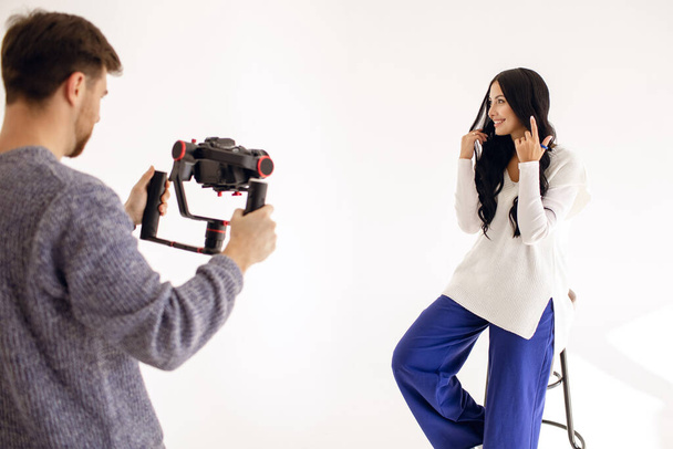 A woman blogger shoots a video blog in the studio with the help of a videographer with a professional video camera. Modern work as a blogger or journalist. The brunette straightens her hair - Photo, Image