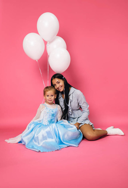 Γοητευτικό χαμογελαστό κορίτσι γενεθλίων με λευκά μπαλόνια και η νεαρή ευτυχισμένη μελαχρινή μητέρα της με μπλε φορέματα κάθονται σε ένα ροζ φόντο. Χρόνια πολλά στη μαμά και την κόρη. - Φωτογραφία, εικόνα