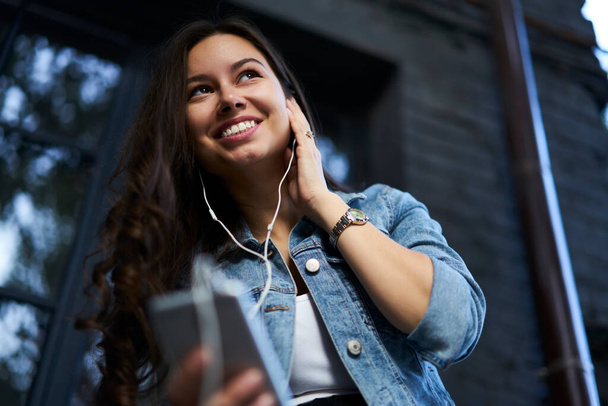 Emotional female user listening funny music in earphones connected to cellphone during recreation time in city, cheerful hipster girl with mobile technology enjoying audio song from playlist - Foto, Imagen