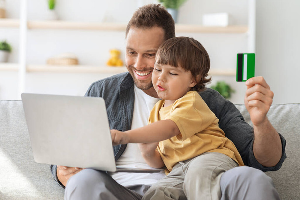 Online shopping fun. Cute little boy making purchases in internet with daddy, choosing good on laptop screen - Photo, Image