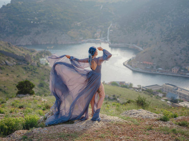 Mulher bonita nova com cabelo azul em vestido cinza olhando para as montanhas do mar. Senhora na natureza. Fotos de moda. Pano de seda acenando no vento - Foto, Imagem