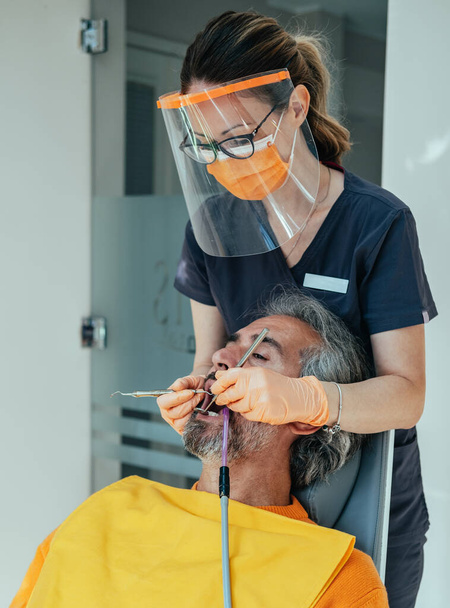 Mužský pacient sedí na zubařské židli s otevřenou pusou a dostává zubní protézu od zubní lékařky v ochranných pracovních oděvech na zubní klinice během COVID-19 - Fotografie, Obrázek
