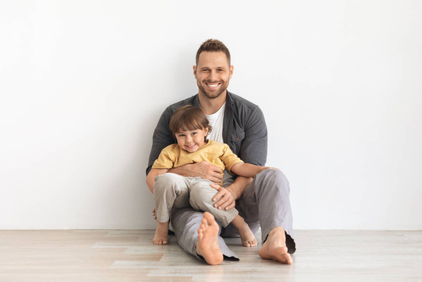Rodzinna miłość i szczęście. Wewnętrzny portret pozytywnego młodego ojca obejmującego swojego synka, siedzącego razem na podłodze - Zdjęcie, obraz