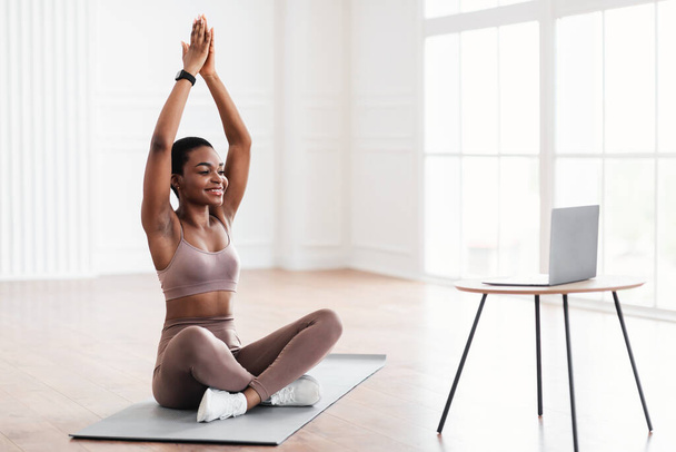 Sonriente joven mujer negra sentada en la esterilla de yoga utilizando el ordenador portátil - Foto, imagen