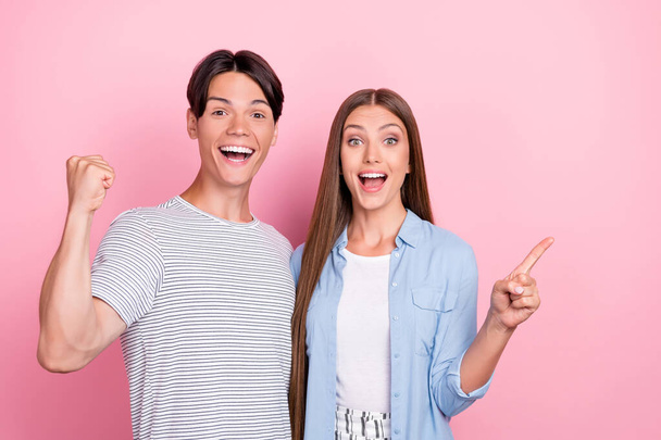 Photo of cute lucky young lovers dressed casual outfits pointing finger empty space rising fist smiling isolated pink color background - Valokuva, kuva