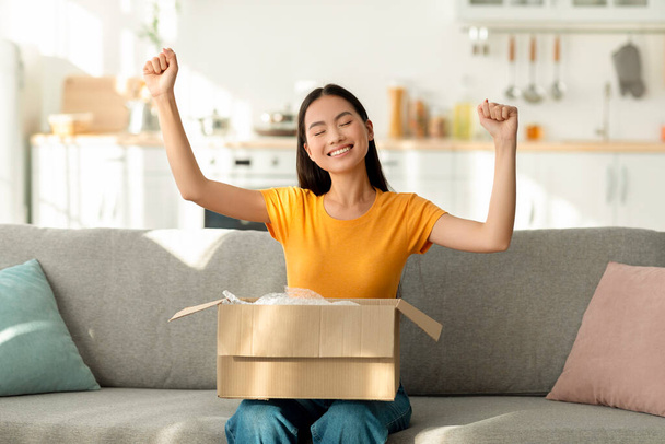 Banner de compras y entrega en línea. Encantado asiático señora abriendo paquete y haciendo SÍ gesto, sentado en el sofá - Foto, imagen