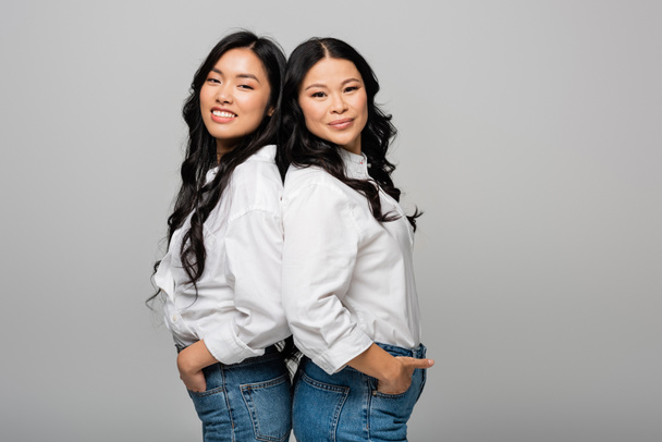felice asiatica madre e figlia in blue jeans e camicie bianche in posa con le mani in tasche isolate su grigio - Foto, immagini
