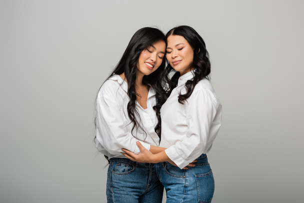 onnellinen aasialainen äiti ja tytär sinisissä farkuissa ja valkoisissa paidoissa halaus eristetty harmaalla - Valokuva, kuva