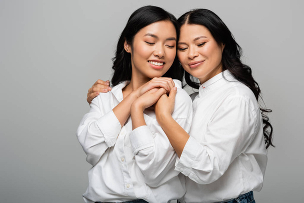 cheerful asian mother and young adult daughter hugging each other and holding hands isolated on grey - Photo, Image