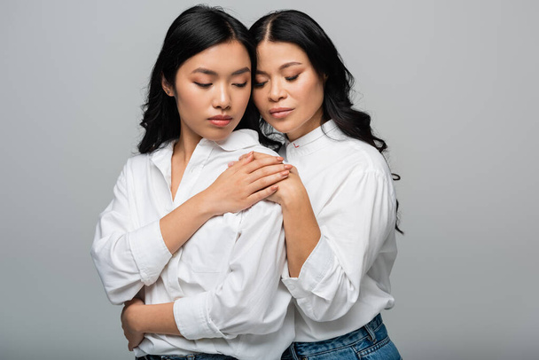 brunette mother and asian young daughter hugging each other and holding hands isolated on grey - Foto, immagini