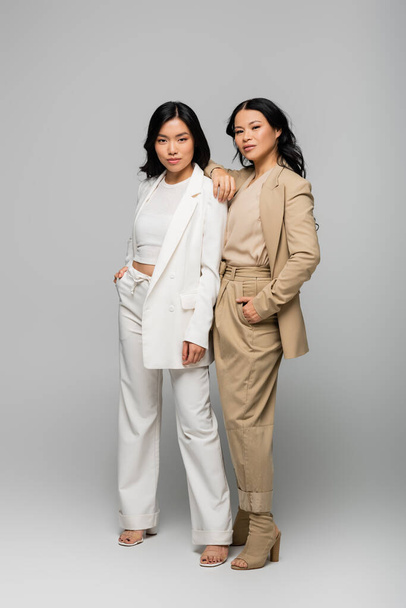 full length of trendy asian mother and young adult daughter in suits posing on grey - Foto, afbeelding