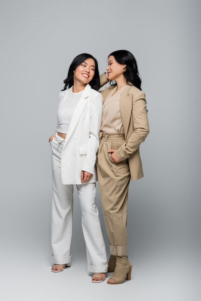 full length of stylish asian mother and young adult daughter in suits smiling on grey - Fotografie, Obrázek