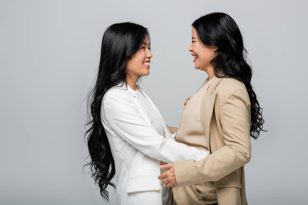stylish asian mother and happy young adult daughter in suit hugging isolated on grey - Photo, Image