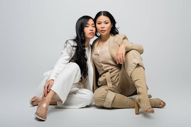 piena lunghezza di elegante asiatica madre in abito e bruna giovane figlia adulta seduta sul grigio - Foto, immagini