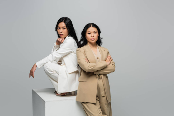 asian young adult woman sitting on cube near brunette mother posing with crossed arms isolated on grey - Fotoğraf, Görsel