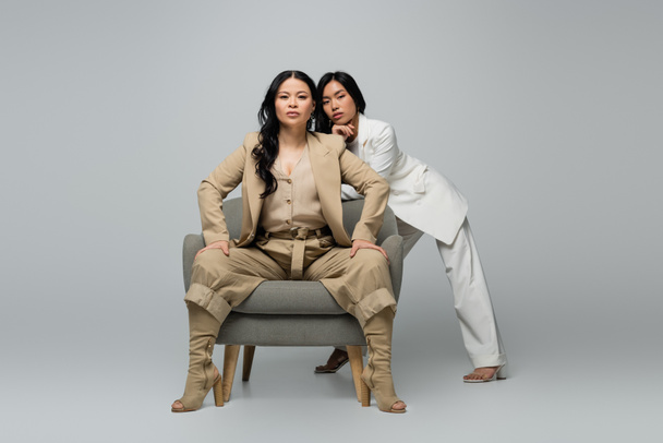 full length of asian woman sitting in armchair near trendy young daughter on grey - Foto, afbeelding