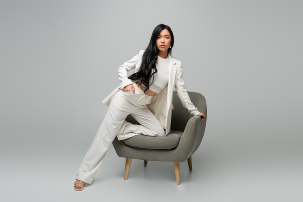 full length of young asian woman leaning on armchair while posing with hand in pocket on grey - Photo, Image