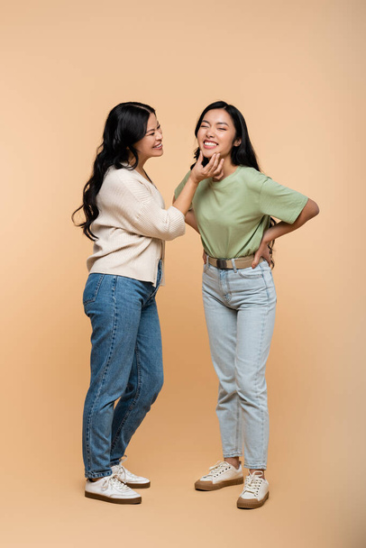 full length of cheerful asian mother touching face of pleased young adult daughter on beige  - Foto, immagini