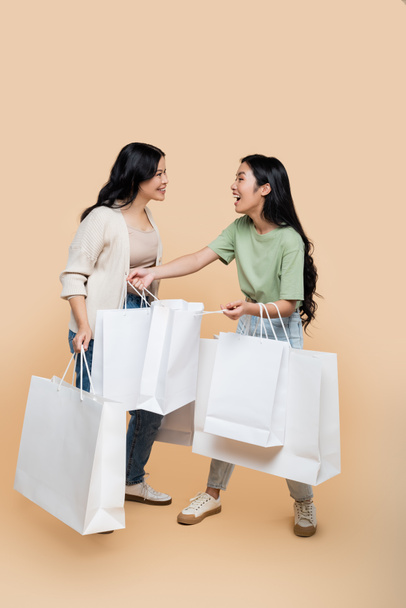 full length of cheerful asian mother and daughter with shopping bags on beige - Foto, Imagem