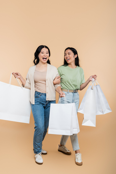 full length of asian mother and young adult daughter smiling while walking with shopping bags on beige  - Photo, image
