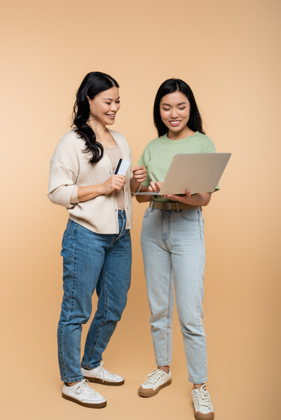 glücklich asiatische Mutter und erwachsene Tochter mit Kreditkarte Blick auf Laptop beim Online-Shopping auf beige  - Foto, Bild