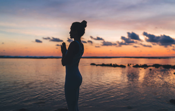 Side silhouette view of calm girl in sportive clothing praying and meditating for getting soul inspiration, woman in namaste thinking about harmony and mindfulness recreating at evening seashore - Fotoğraf, Görsel