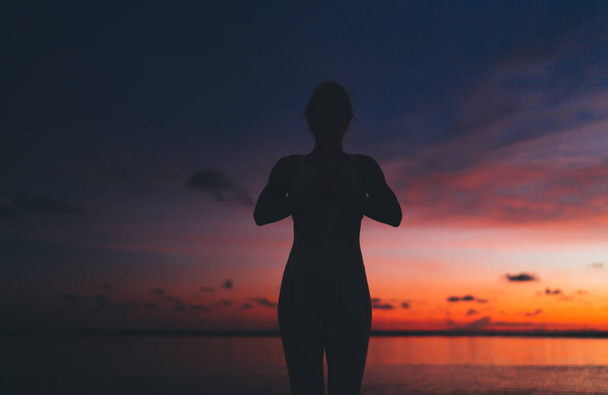 Silhouette of fit female practicing yoga standing on Bali beach in Anjali Mudra posture with pink and orange sunset sky behind - Foto, Bild