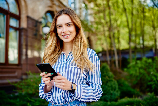 Young joyful female with long fair hair hair using smartphone and looking at camera while standing on blurred background of street - Фото, изображение