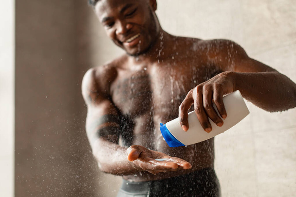 beau noir guy prendre douche serrant shampooing bouteille dans salle de bain - Photo, image