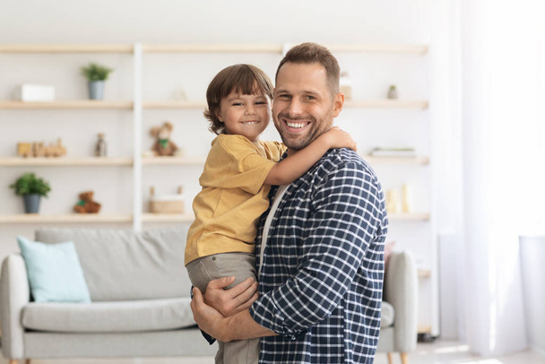 Família feliz. Interior retrato de homem caucasiano positivo abraçando seu filho bonito, segurando-o em mãos - Foto, Imagem