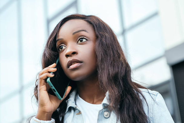 Sérieux belle afro-américaine jolie femme tenir la main téléphone parler bavarder amis collègue debout rue dehors conversation parler fond flou bokeh - Photo, image