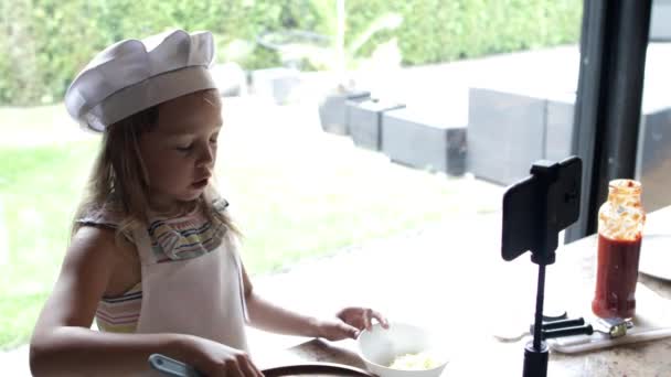 Маленькая девочка готовит пиццу и снимает видео на смартфоне для видеоблога на домашней кухне - Кадры, видео