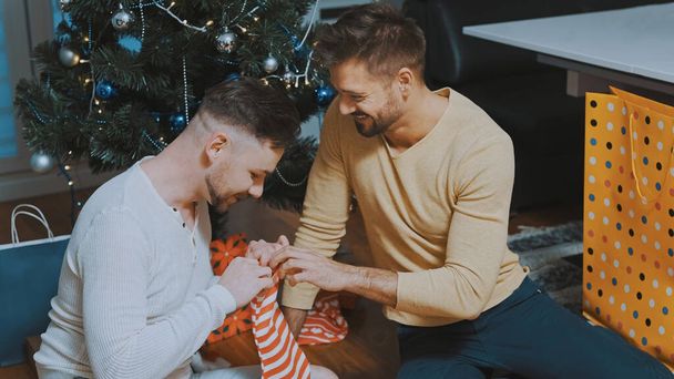 Young guy male couple gifting each other with christmas stockings. Homosexual relationship and winter holidays concept - Foto, Bild