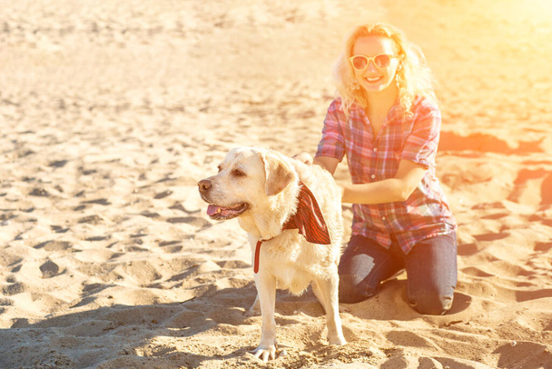 Портрет молодой красивой женщины в солнечных очках, сидящей на песчаном пляже с золотой собакой-ретривером. Девушка с собакой на море. Солнечная вспышка - Фото, изображение