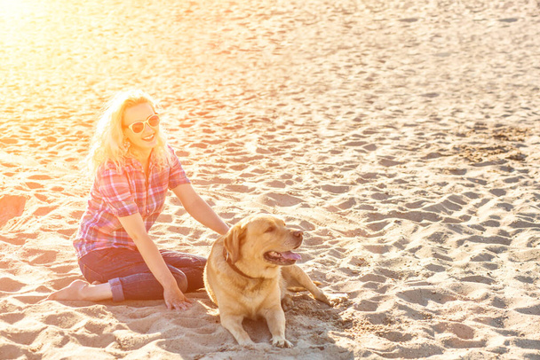 Portrait of young beautiful woman in sunglasses sitting on sand beach with golden retriever dog. Girl with dog by sea. Sun flare - Fotó, kép