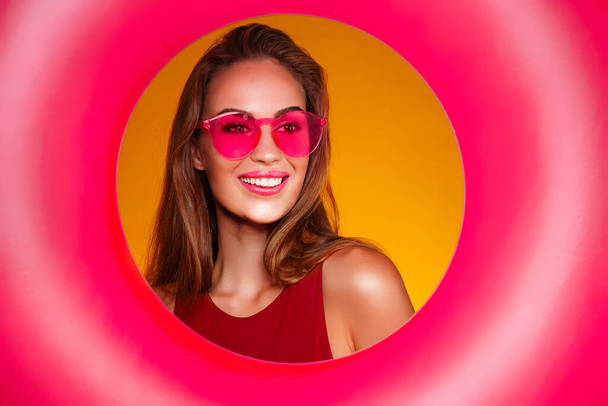 Portrait of cheerful young woman in pink sunglasses who holds inflatable ring over yellow background - Φωτογραφία, εικόνα