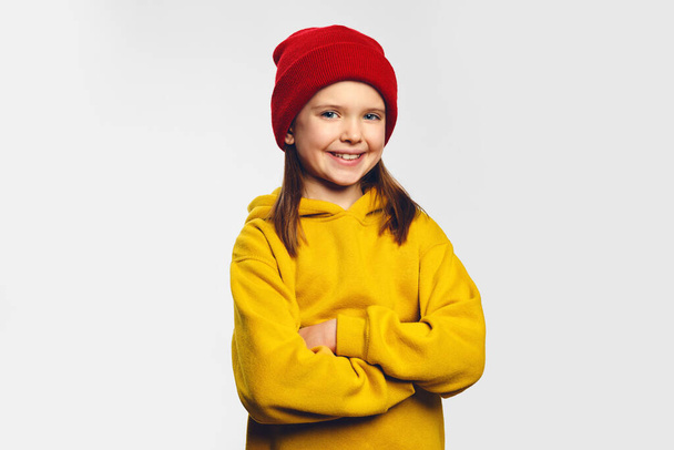 Hipster kid in yellow hoodie and red hat holds hands crossed and smiles broadly - Foto, Imagem