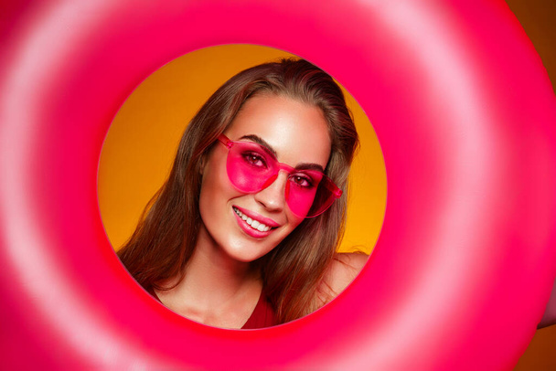 Portrait of young woman in pink sunglasses who holds inflatable ring over yellow color background - Foto, Imagen