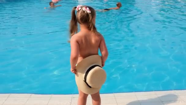 divertida niña de 3 años baila y posa cerca de los niños nadadores cerca de la piscina, vista trasera. Infancia, relación, concepto de vacaciones de verano. enfoque selectivo en el sombrero - Metraje, vídeo
