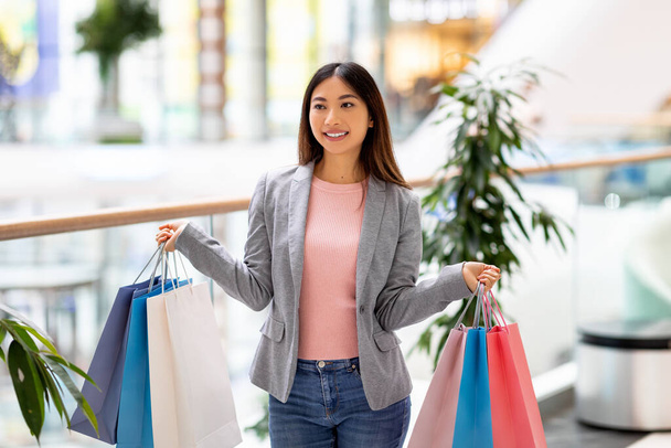 Porträt der schönen jungen Asiatin mit bunten Geschenktüten in beiden Händen beim Einkaufen in der City Mall - Foto, Bild