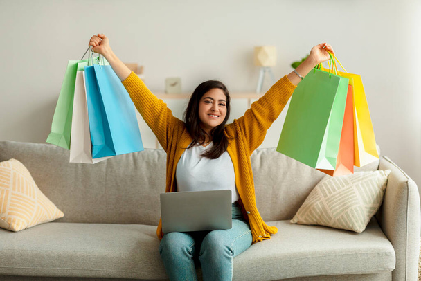 Alegre árabe señora de compras en línea, sentado en el sofá con el ordenador portátil, levantando bolsas de regalo por encima de su cabeza en la emoción - Foto, Imagen