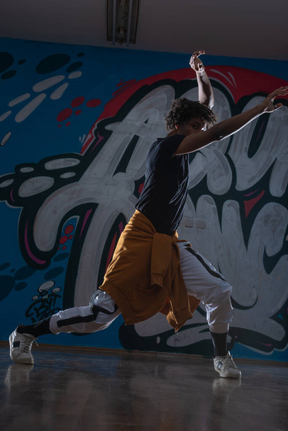 Młody czarny mężczyzna taniec hip hop styl w środowisku miejskim. ma na sobie pomarańczowy strój i jest na tle graffiti. - Zdjęcie, obraz