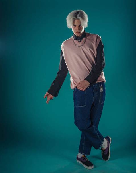 Young adult man in casual outfit wear looking at camera, holding skateboard near face and standing at studio on blue background - Foto, immagini