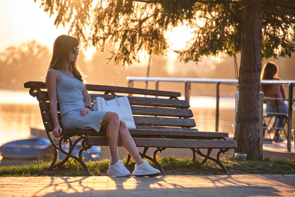 Gölün kıyısındaki bankta tek başına oturan yalnız genç kadın sıcak yaz akşamının tadını çıkarıyor. Doğada refah ve rahatlama kavramı. - Fotoğraf, Görsel