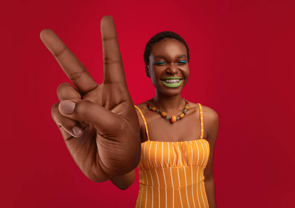 Щаслива чорна жінка з яскравим макіяжем у стильному вбранні, що демонструє велику перемогу або жест миру на червоному тлі
 - Фото, зображення