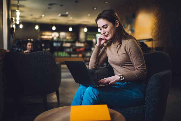 Positive female in casual wear sitting on armchair in creative workspace and browsing laptop while messaging with friends and waiting for order - Photo, Image
