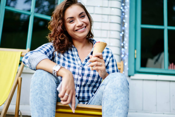 Portrait of funny hipster girl smiling at camera during leisure for eating delicious sorbet ice cream in cone waffle, happy millennial woman with gelato dessert posing outdoors while resting - Foto, imagen
