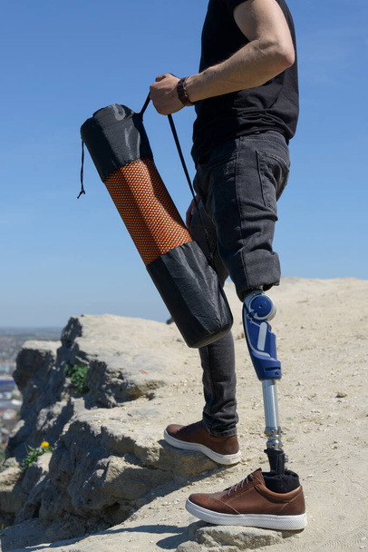 Ένας άντρας με προσθετικό πόδι ταξιδεύει στα βουνά. Ντυμένος με μαύρο τζιν και μπλουζάκι, κουβαλάει χαλάκι. - Φωτογραφία, εικόνα